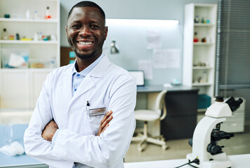 Naklejka na ściany i meble Waist up portrait of black young man wearing lab coat posing in clinic laboratory and smiling, copy space