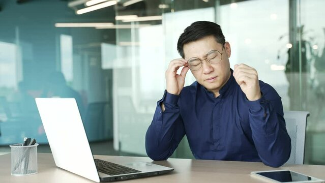 Tired overworked asian entrepreneur business man IT programmer with eyes hurt massaging having bad poor vision after hard day working on computer typing and browsing at desk in modern office indoors 