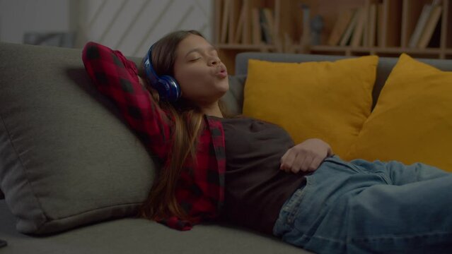Joyful cute Hispanic teenage girl with eyes closed in wireless headphones relaxing on sofa, enjoying music and singing along to catching song while spending leisure in domestic room.