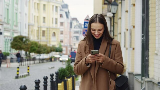 Cheerful woman writing sms with smartphone. Dreamy stylish female chatting at street. Blogger subscribing new social media, buying in internet, ordering products online. Mobile addiction concept