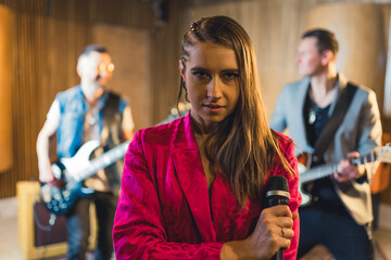 Focused serious stylish female vocalist looking straight to the camera. Blurred male guitarists in the background. Recording studio concept. High quality photo