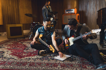 Caucasian guitarist sitting on patterned carpet and discussing their new songs. Electric guitars in...