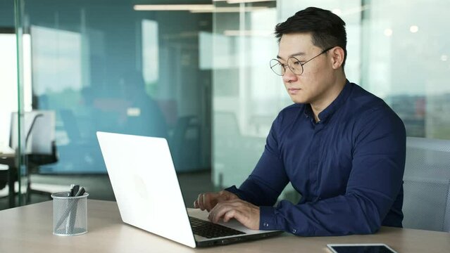 Busy IT specialist programmer with glasses working typing coding on laptop computer in modern office centre Asian entrepreneur professional developer thinking analyzing finance digital data indoors