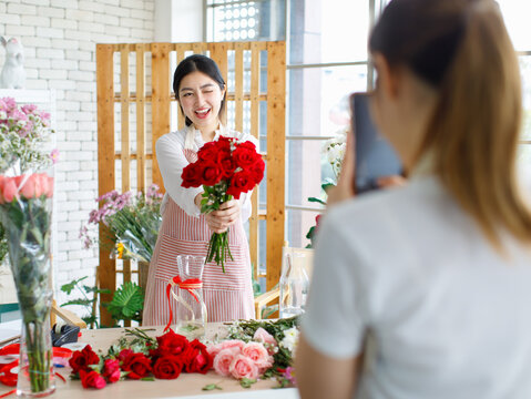 Millennial Asian young beautiful professional female flower shopkeeper owner decorator florist wearing apron standing smiling holding red roses bunch bouquet while unrecognizable colleague take photo