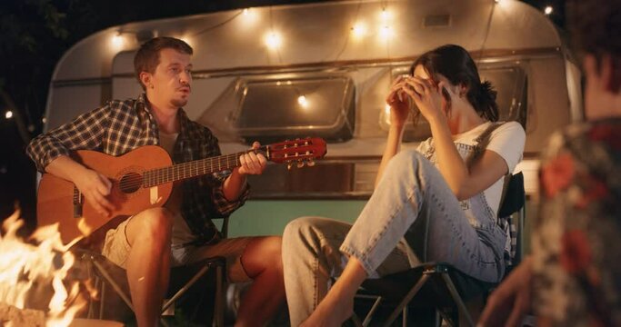 Beautiful Young Friends Enjoy a Relaxing Summertime Evening at a Camping Rest Area. Singing Songs, Playing Guitar, Dancing at a Campfire Next to Their Caravan with Decorative Lights.