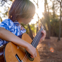 Boy plays the guitar in the park sitting on a bench.  Child plays a musical instrument, in nature. ...