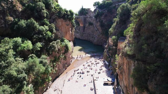 A cinematic aerial shot moving forward between steep cliffs towards the historic bridge at the picturesque Fiordo di Furore, a scenic vacation destination for tourists on the Amalfi Coast in Italy.