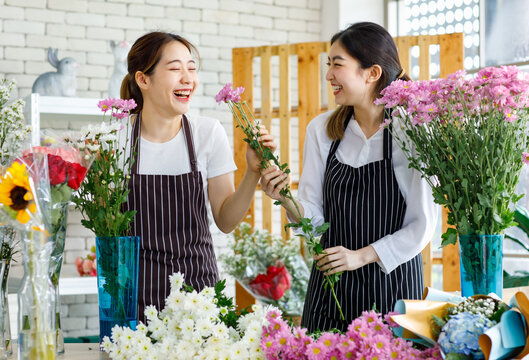 Millennial Asian young female flower shopkeeper decorator florist worker in apron smiling holding white flower bunch bouquet colleague use scissors cutting arranging decorating stalk in floral store