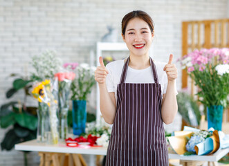 Portrait studio shot of Millennial Asian young female flower shopkeeper decorator florist employee worker in apron standing smiling holding thumbs up in front blurred workshop table in floral store