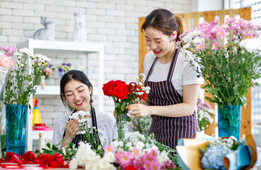 Fototapeta premium Millennial Asian young female flower shopkeeper decorator florist worker in apron smiling holding flower helping bunch bouquet colleague arranging decorating stalk in floral store.