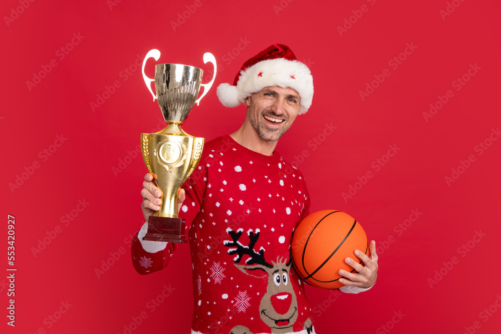 Wall mural Santa trainer hold basketball ball and champion cup. Portrait of middle aged man in sweater isolated over red background. Concept of holidays, happiness, emotions and Christmas celebration.