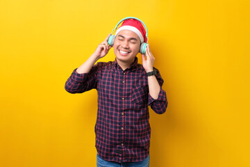 Happy young Asian man in Santa hat listening music with headphones, standing with closed eyes on yellow studio background. Happy New Year 2023 celebration merry holiday concept