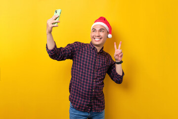 Fototapeta na wymiar Cheerful young Asian man in Santa hat taking selfie on smartphone, showing victory sign over yellow studio background. Happy New Year 2023 celebration merry holiday concept