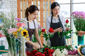 Millennial Asian young female flower shopkeeper decorator florist worker in apron smiling holding flower bunch bouquet colleague arranging decorating stalk in floral store.