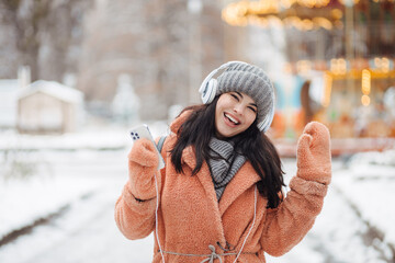 Pretty young long-haired woman in pink coat enjoy winter day and listening music with headphones at snowy park