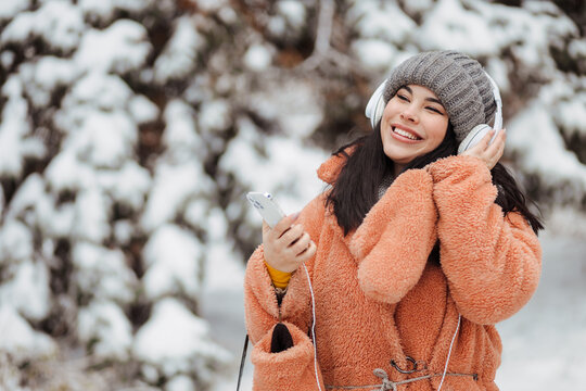 Pretty Young Long-haired Woman In Pink Coat Enjoy Winter Day And Listening Music With Headphones At Snowy Park