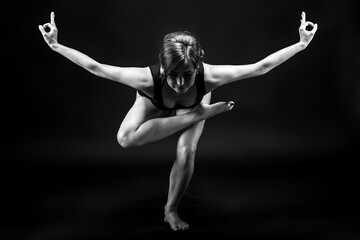 A young, slender, athletic, red-haired girl in a black T-shirt and black leggings sits in an asana on a black background in a photo studio.