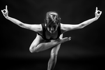 A young, slender, athletic, red-haired girl in a black T-shirt and black leggings sits in an asana on a black background in a photo studio.