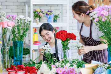 Millennial Asian young female flower shopkeeper decorator florist worker in apron smiling holding flower helping bunch bouquet colleague arranging decorating stalk in floral store.