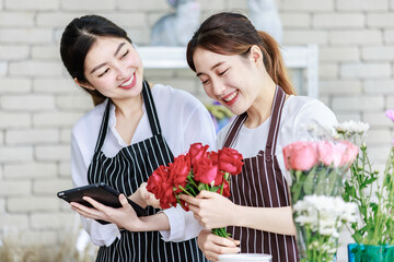 Millennial Asian young professional flower shopkeeper florist employee worker wearing apron standing smiling holding red roses bunch bouquet while colleague using tablet computer in floral store