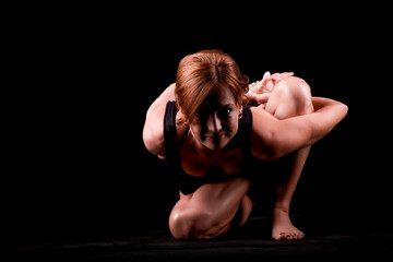 Fototapeta na wymiar A young, slender, athletic, red-haired girl in a black T-shirt and black leggings sits in an asana on a black background in a photo studio.