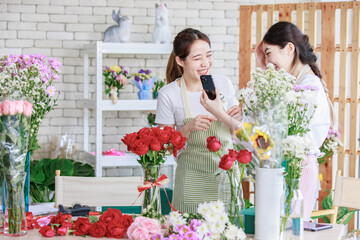 Millennial two Asian young professional female flower shopkeeper owner decorator wearing apron standing smiling using smartphone taking photo of decorating red roses bunch bouquet in vase in store