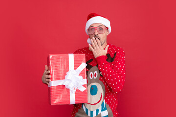 surprised man in winter sweater and santa claus hat. xmas guy with present box on red background.