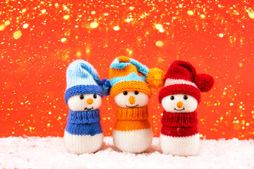 Knitted snowmen in a red, blue and orange hats and sweater stands on a red background with golden Christmas lights. Merry Christmas and New Year 2023 greeting card