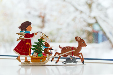 Christmas swiss traditional metal toy angel girl in red dress with presents rides on sleigh with reindeer and rabbit against blurred winter window background - Powered by Adobe