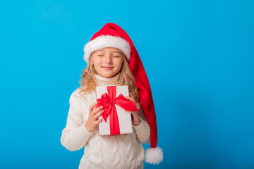 A cute little girl in a Santa hat holds a gift on a blue background in the studio. Space for text. Winter concept, knitted sweater