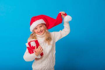 A cute little girl in a Santa hat holds a gift on a blue background in the studio. Space for text. Winter concept, knitted sweater