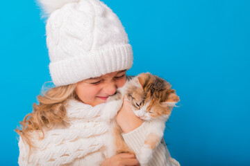 Obraz premium a cute little girl in a white knitted hat and sweater is holding a kitten on a blue background in the studio. Space for text. The concept of Christmas, the symbol of the year