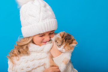 a cute little girl in a white knitted hat and sweater is holding a kitten on a blue background in the studio. Space for text. The concept of Christmas, the symbol of the year