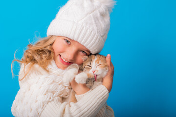 Obraz premium a cute little girl in a white knitted hat and sweater is holding a kitten on a blue background in the studio. Space for text. The concept of Christmas, the symbol of the year