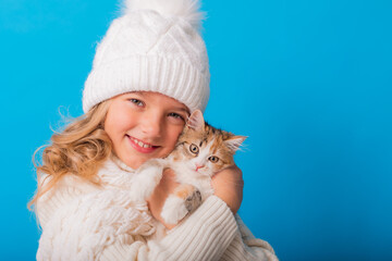 Obraz premium a cute little girl in a white knitted hat and sweater is holding a kitten on a blue background in the studio. Space for text. The concept of Christmas, the symbol of the year