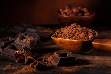 in the foreground, in a dark environment, cocoa powder, pieces of dark chocolate and hazelnuts - obrazy, fototapety, plakaty