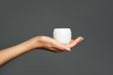 Skincare beauty product. Studio photo of female hand is holding a white jar with cream against grey background. Daily care and hygiene routine.