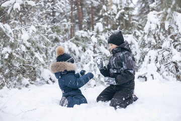 Fototapeta na wymiar Happy teenager and little boy playing in snow in winter forest. Children having fun snowball fight together outdoors. Christmas holidays and New year family vacation. Brothers walking in park