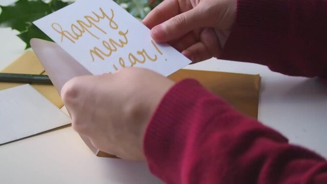 Closeup of a person at desk, opening mail during the winter holidays: a golden envelope containing a greeting card wishing a happy new year in handwritten, gold calligraphy