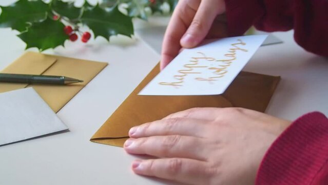 Closeup of the hands of a woman at her desk, opening a golden envelope from friends or family containing a handwritten card wishing happy holidays, on Christmas day or during the winter festive season