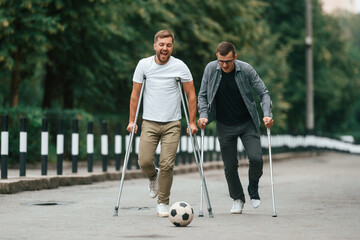 Playing with soccer ball. Two men with crutches is outdoors on the road