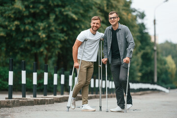 Standing and smiling. Two men with crutches is outdoors on the road
