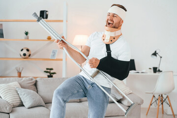 Standing, holding crutch and pretending that he's playing guitar. Having fun. Man is at home...