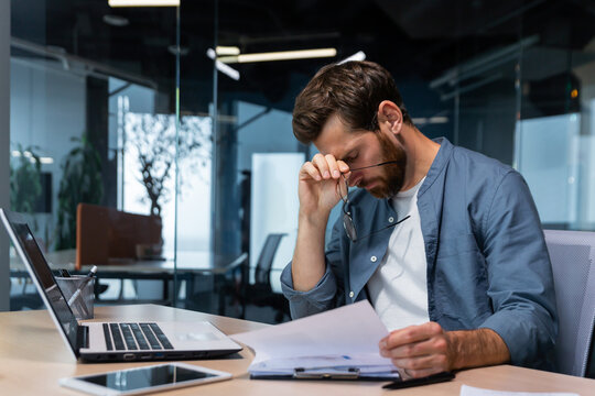 Upset businessman behind paper work inside modern office, mature man with beard reading financial reports and account documents unhappy with results and disappointed with achievements.