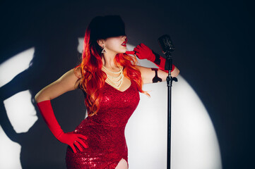Pretty woman singer or movie actor in red dress with red hair holding microphone on a stage. Star singer on stage