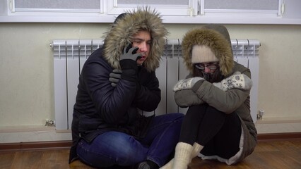 A young couple is sitting on the floor by the radiator in winter jackets. A man calls on the phone in a company. to turn on the heating.