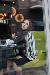 Obraz na płótnie Canvas A beautiful young woman in a black dress sits in a cafe by the window and drinks coffee. Coffee break