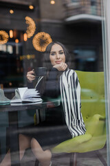 A beautiful young woman in a black dress sits in a cafe by the window and drinks coffee. Coffee break