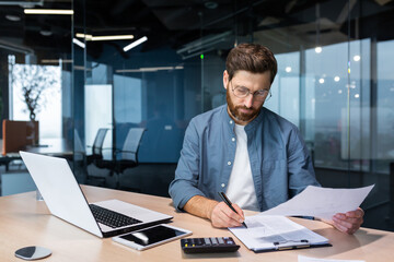 Serious and focused financier accountant on paper work inside office, mature man using calculator and laptop for calculating reports and summarizing accounts, businessman at work in casual clothes.