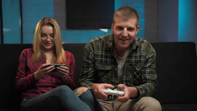 Man playing video games at concole while his girl play game on smartphone in their living room.Young Guy using console gamepad looking at TV having fun sitting on the couch at home.Gambling addiction 